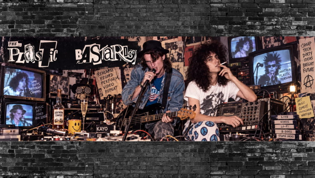 The Plastic Bastards surrounded by old TVs, tape decks and cassettes, one singing into a microphone and one smoking beside a mixing desk